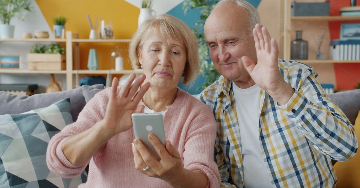 Un couple de personnes âgées utilise son smartphone pour une conversation vidéo et se salue de la main.