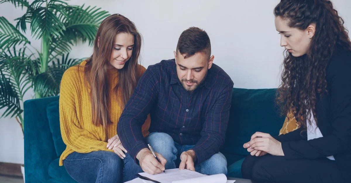 Jeune couple consulte un conseiller financier, signant des documents importants à l'intérieur.