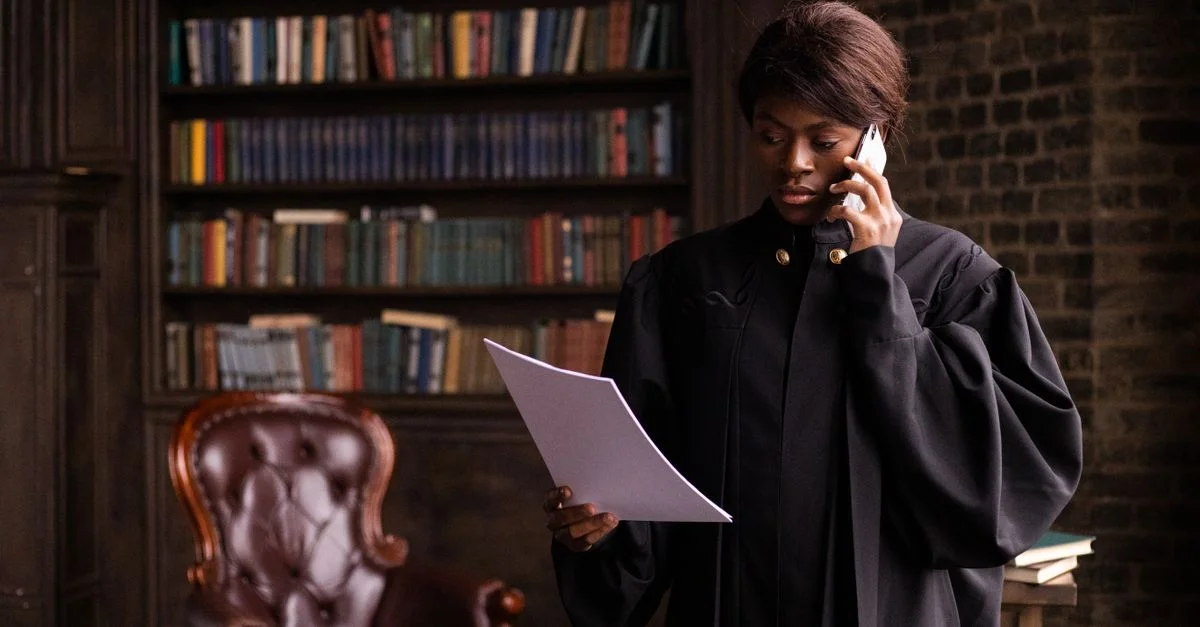 Un juge afro-américain en robe traditionnelle tient un document et parle au téléphone dans une bibliothèque de droit.